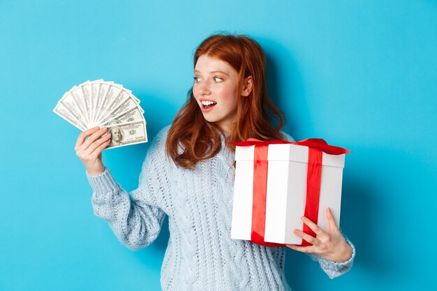 Weihnachts- und Einkaufskonzept. Aufgeregtes rothaariges Mädchen, das Dollar betrachtet, großes Neujahrsgeschenk hält, Geschenke kauft und auf blauem Hintergrund steht
