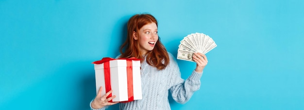 Weihnachts- und Einkaufskonzept begeisterte rothaarige Mädchen, die Dollars betrachten, die einen großen Geschenkkauf für das neue Jahr halten