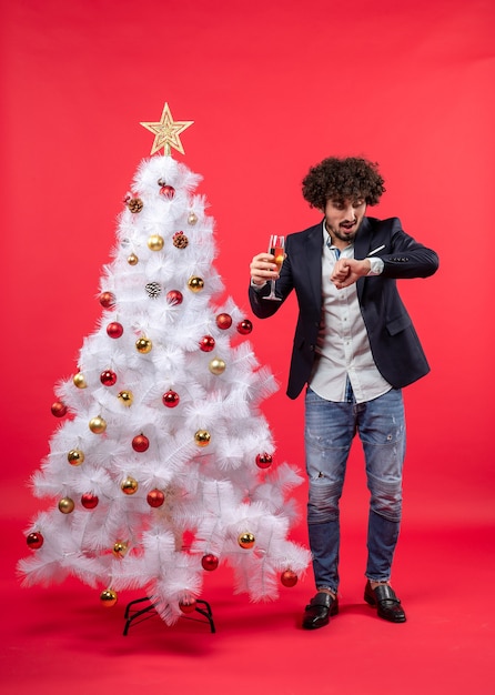Weihnachtsfeier mit bärtigem jungen mann mit weinprüfzeit und stehend nahe weihnachtsbaum auf rot Kostenlose Fotos