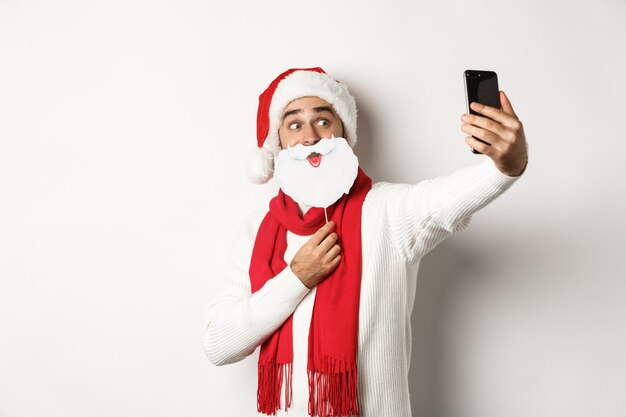 Weihnachtsfeier und Feierkonzept. Junger Mann, der Selfie mit lustiger weißer Bart-Sankt-Maske und -Hut macht und für Fotos auf dem Handy posiert, Studiohintergrund