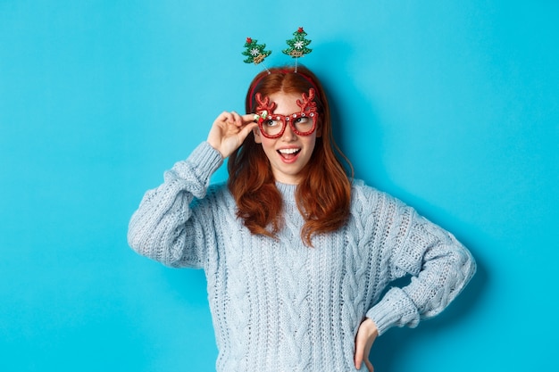 Weihnachtsfeier und Feierkonzept. Süßes rothaariges Teenie-Mädchen, das Neujahr feiert, Weihnachtsbaum-Stirnband und lustige Brille trägt, amüsiert nach links schaut, blauer Hintergrund