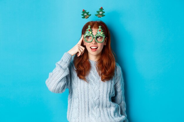 Weihnachtsfeier und Feierkonzept. Süßes rothaariges Teenie-Mädchen, das Neujahr feiert, Weihnachtsbaum-Stirnband und lustige Brille trägt, amüsiert nach links schaut, blauer Hintergrund