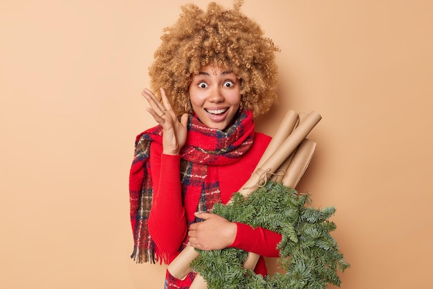 Weihnachtsfeiern Konzept. Aufgeregt fröhliche junge Frau hebt die Hand, um den Weihnachtskranz zu dekorieren, der Floristendekorateur ist und einen Schal um den Hals trägt, der auf beigem Hintergrund isoliert ist.