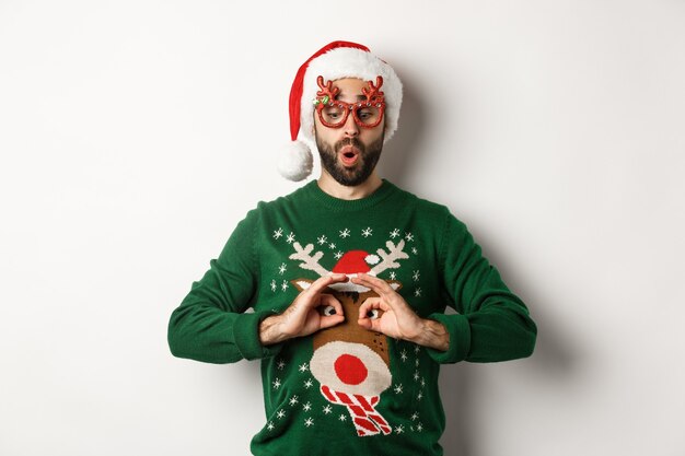 Weihnachtsferien, Feierkonzept. Fröhlicher Kerl in Weihnachtsmütze und Partybrille, der sich über einen lustigen Pullover lustig macht und auf weißem Hintergrund steht