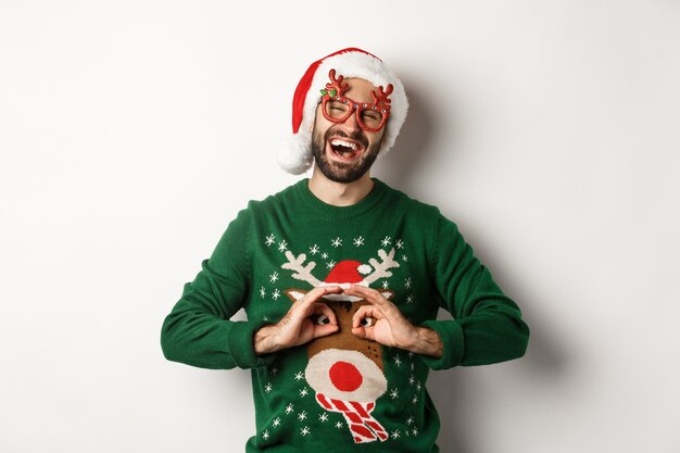 Weihnachtsferien, Feierkonzept. Fröhlicher Kerl in Weihnachtsmütze und Partybrille, der sich über einen lustigen Pullover lustig macht und auf weißem Hintergrund steht