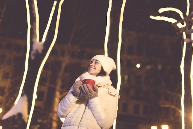 Weihnachtsferien. schöne lächelnde frau in warmer kleidung mit tasse heißem kaffee im freien im winter. stadt nacht.