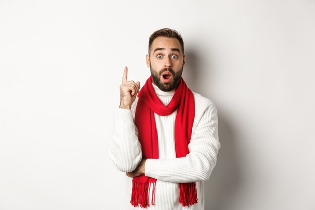 Weihnachtsferien und Feierkonzept. Aufgeregter bärtiger Mann, der eine Idee hat, den Finger hebt und einen Plan vorschlägt, in rotem Schal mit Pullover, weißem Hintergrund steht.