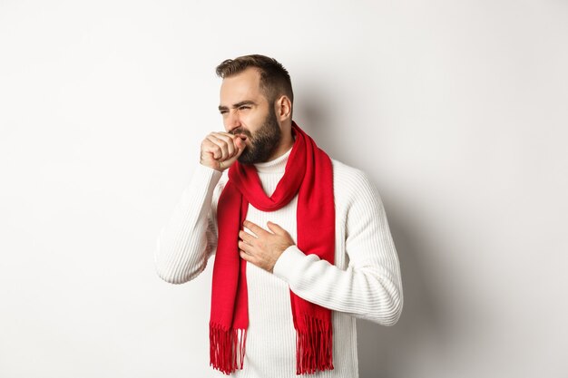 Weihnachtsferien und Feierkonzept. Mann fühlt sich krank, hustet und verzieht das Gesicht von Halsschmerzen, Covid-19-Symptome an Silvester, stehend auf weißem Hintergrund.