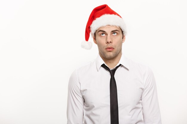 Weihnachtskonzept Gutaussehender Geschäftsmann trägt eine Weihnachtsmütze, die mit nachdenklichem Gesichtsausdruck auf weißem, isoliertem Hintergrund posiert