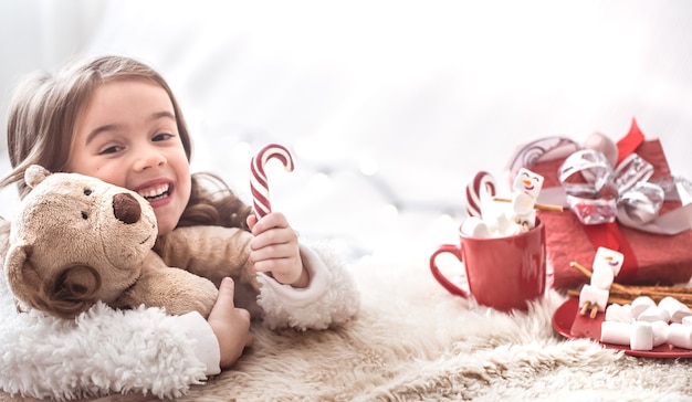 Weihnachtskonzept, kleines niedliches Mädchen, das Teddybärspielzeug im Wohnzimmer mit Geschenken auf hellem Hintergrund umarmt, Platz für Text