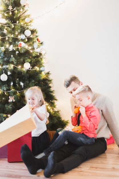 Weihnachtskonzept mit dem Vater und Kindern, die nahe bei Weihnachtsbaum sitzen