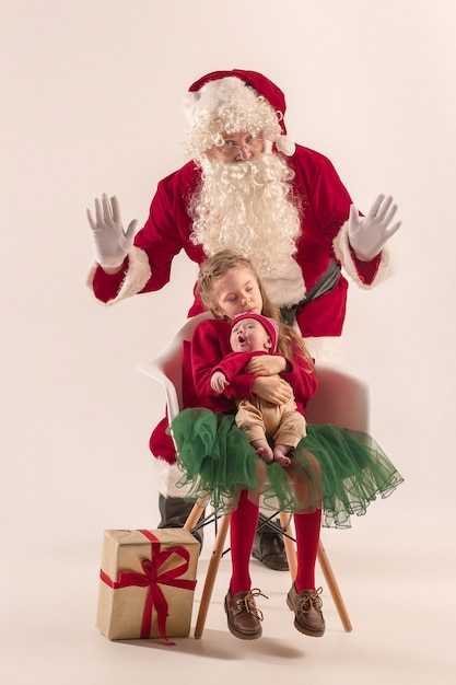 Weihnachtsporträt des niedlichen kleinen neugeborenen Mädchens, der hübschen jugendlich Schwester, gekleidet in Weihnachtskleidung und Mann, der Weihnachtsmannkostüm und -hut, Studioaufnahme, Winterzeit trägt. Das Weihnachts-, Feiertagskonzept