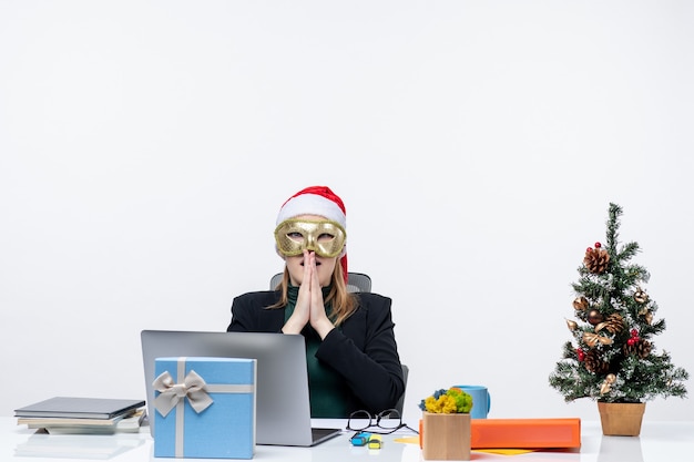 Weihnachtsstimmung mit angespannter junger Frau mit Weihnachtsmannhut und tragender Maske, die an einem Tisch auf einem weißen Hintergrund sitzt