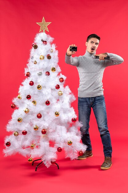 Weihnachtsstimmung mit dem jungen Mann, der nahe geschmücktem Weihnachtsbaum steht und Mikrofon hält und sein Foto macht