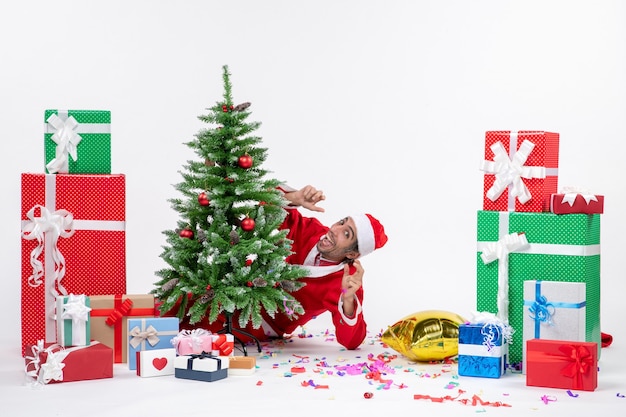 Weihnachtsstimmung mit dem jungen Weihnachtsmann, der sich hinter Weihnachtsbaum nahe Geschenken in verschiedenen Farben auf weißem Hintergrund versteckt zeigt