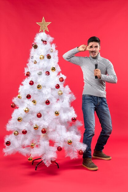 Weihnachtsstimmung mit dem selbstbewussten Kerl, der in Jeans gekleidet ist, die nahe geschmücktem Weihnachtsbaum stehen und Mikrofon halten und lustige Gesten zeigen