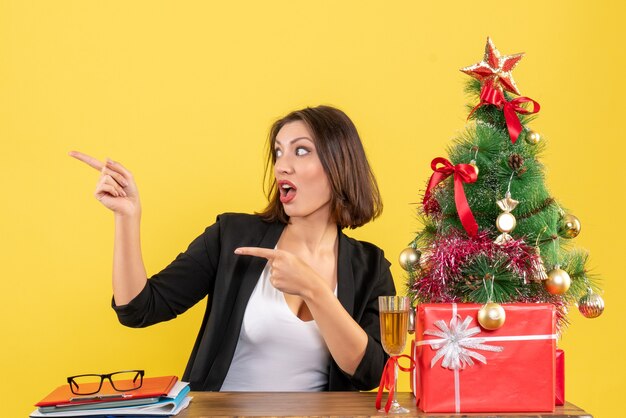 Weihnachtsstimmung mit der jungen angespannten Geschäftsdame, die jemanden zeigt und im Büro auf Gelb sitzt