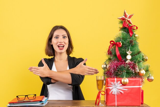 Weihnachtsstimmung mit der jungen zufriedenen glücklichen emotionalen Geschäftsdame, die etwas zeigt und an einem Tisch im Büro auf Gelb sitzt