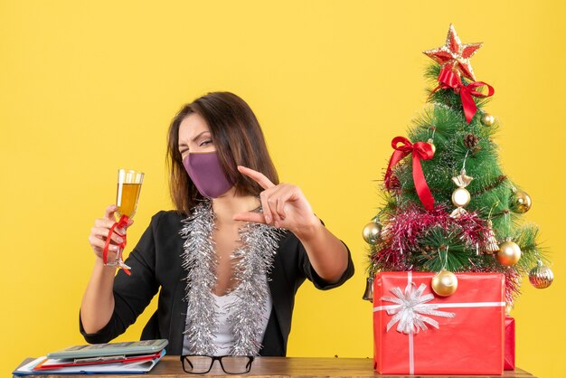 Weihnachtsstimmung mit der schönen Dame im Anzug, die medizinische Maske trägt und Wein im Büro auf Gelb züchtet