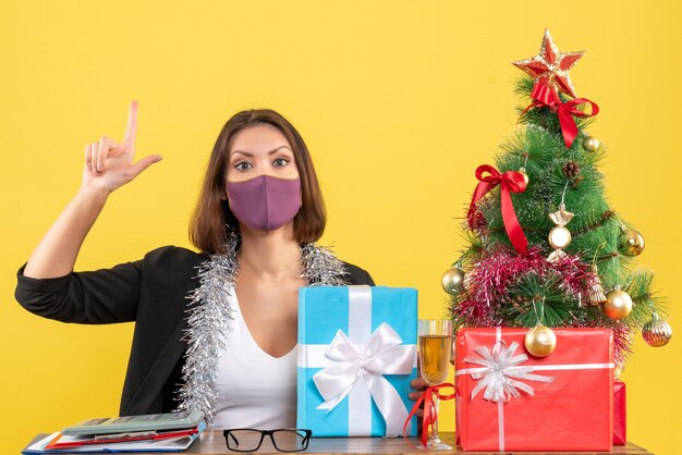 Weihnachtsstimmung mit der schönen Dame im Anzug mit der medizinischen Maske und im Halten des Geschenks, das oben im Büro auf Gelb zeigt