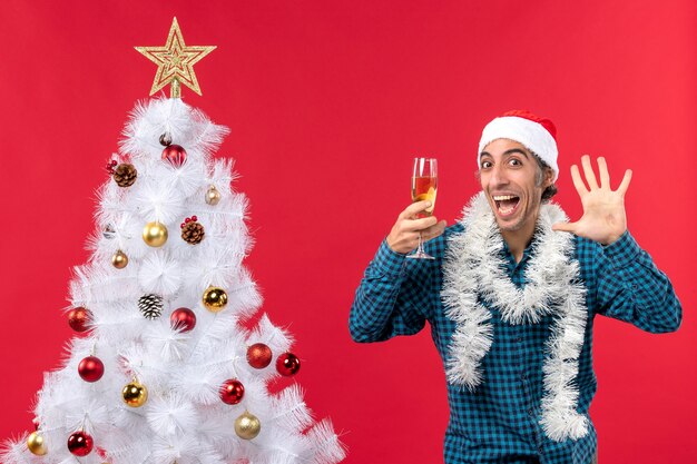 Weihnachtsstimmung mit emotionalem jungem Mann mit Weihnachtsmannhut in einem blauen gestreiften Hemd, das ein Glas Wein hält und fünf nahe Weihnachtsbaum zeigt