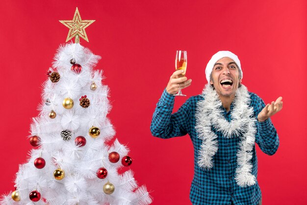 Weihnachtsstimmung mit emotionalem jungem Mann mit Weihnachtsmannhut in einem blauen gestreiften Hemd, das ein Glas Wein hält und nahe Weihnachtsbaum lacht