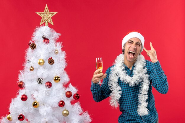 Weihnachtsstimmung mit emotionalem jungem Mann mit Weihnachtsmannhut in einem blauen gestreiften Hemd, das ein Glas Wein nahe Weihnachtsbaum hält