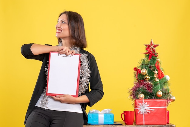 Weihnachtsstimmung mit emotionaler schöner Dame, die im Büro steht und Dokumente hält, die etwas im Büro auf Gelb betrachten