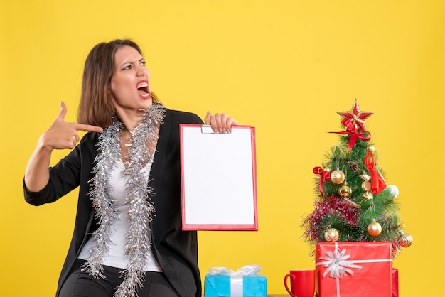 Weihnachtsstimmung mit emotionaler schöner Dame, die im Büro steht und Dokumente im Büro auf Gelb zeigt