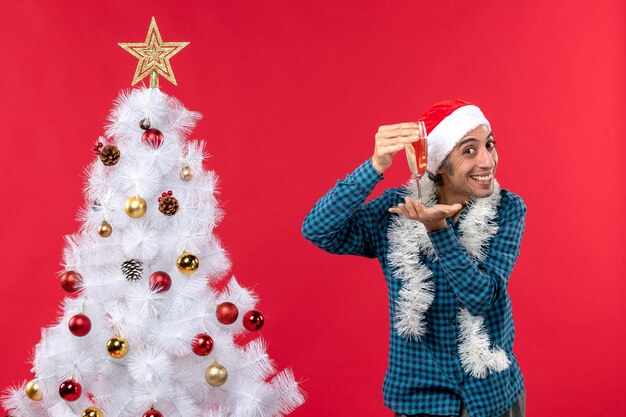 Weihnachtsstimmung mit glücklichem jungem Mann mit Weihnachtsmannhut in einem blauen gestreiften Hemd, das ein Glas Wein nahe Weihnachtsbaum erhebt