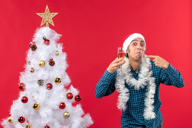 Weihnachtsstimmung mit glücklichem verrückten emotionalen jungen Mann mit Weihnachtsmannhut in einem blauen gestreiften Hemd, das ein Glas Wein erhebt, das sich nahe Weihnachtsbaum zeigt