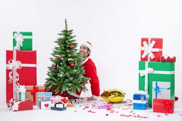 Weihnachtsstimmung mit jungem positivem lustigem Weihnachtsmann, der hinter Weihnachtsbaum nahe Geschenken in verschiedenen Farben auf weißem Hintergrund liegt