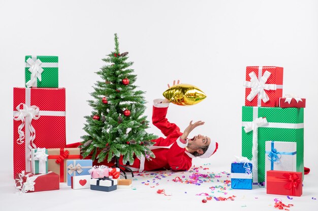 Weihnachtsstimmung mit jungem Weihnachtsmann, der hinter Weihnachtsbaum nahe Geschenken in verschiedenen Farben auf weißem Hintergrund liegt