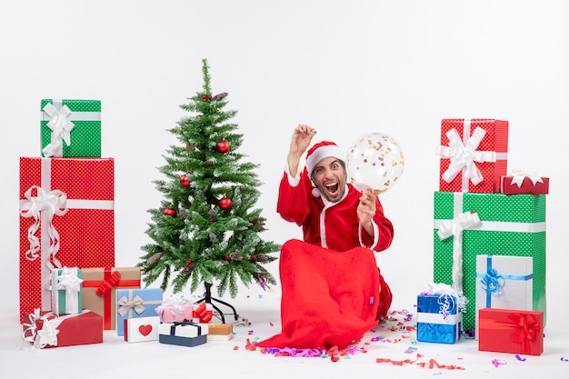 Weihnachtsstimmung mit jungen glücklichen verrückten ha Weihnachtsmann, der nahe Weihnachtsbaum und Geschenken in verschiedenen Farben auf weißem Hintergrund sitzt