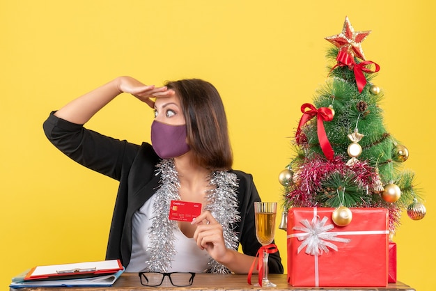 Weihnachtsstimmung mit konzentrierter charmanter dame im anzug, die medizinische maske hält, die bankkarte im büro auf gelb hält