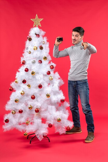 Weihnachtsstimmung mit lächelndem jungen Mann, der nahe geschmücktem Weihnachtsbaum steht und Mikrofon und Telefon hält, die jemanden zeigen, der nach vorne steht