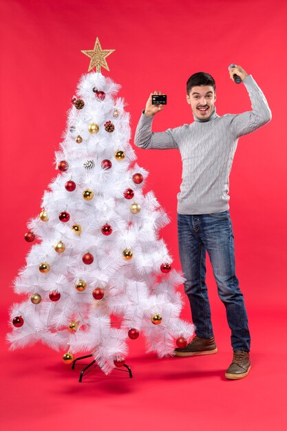 Weihnachtsstimmung mit lächelndem jungen Mann, der nahe geschmücktem Weihnachtsbaum steht und Mikrofon und Telefon hält, die seine Macht zeigen