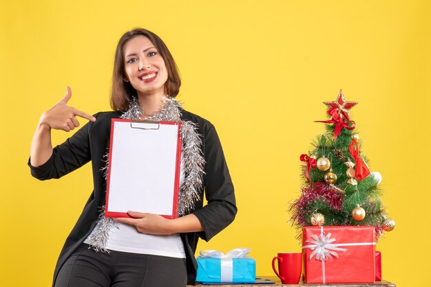 Weihnachtsstimmung mit lächelnder schöner Dame, die im Büro steht und Dokumente im Büro auf Gelb zeigt