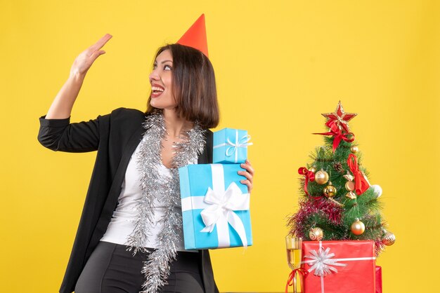 Weihnachtsstimmung mit lächelnder schöner Dame mit Weihnachtshut, der Geschenke hält, die im Büro auf Gelb begrüßen