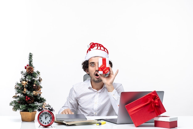 Weihnachtsstimmung mit lustiger Geschäftsperson mit Weihnachtsmannhut, der sein Geschenk zu seinem Gesicht auf weißem Hintergrund erhebt