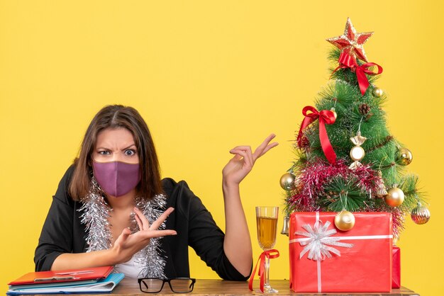 Weihnachtsstimmung mit neugieriger charmanter Dame im Anzug, die medizinische Maske im Büro auf gelbem lokalisiert trägt