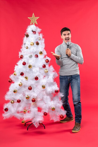 Weihnachtsstimmung mit positivem Kerl gekleidet in Jeans, die nahe geschmücktem Weihnachtsbaum stehen und jemanden anrufen