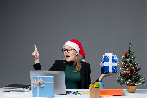 Weihnachtsstimmung mit positiver junger Frau mit Weihnachtsmannhut und Tragen von Brillen, die an einem Tisch sitzen, der Geschenk zeigt, das oben auf dunklem Hintergrund zeigt