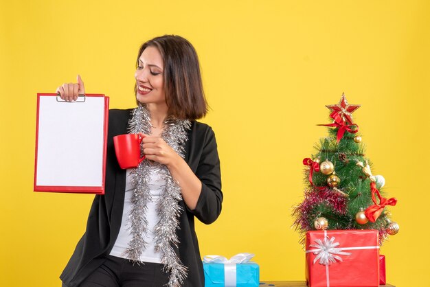 Weihnachtsstimmung mit positiver schöner Dame, die im Büro steht und Dokumentenbecher im Büro auf Gelb hält