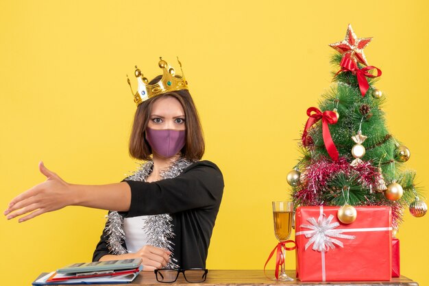 Weihnachtsstimmung mit schöner Dame im Anzug mit medizinischer Maske und tragender Maske, die jemanden im Büro auf Gelb begrüßt