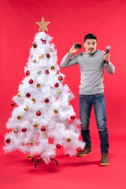 Weihnachtsstimmung mit selbstbewusstem Mann in Jeans, der in der Nähe des geschmückten Weihnachtsbaums steht und Mikrofon und sein Telefon hält