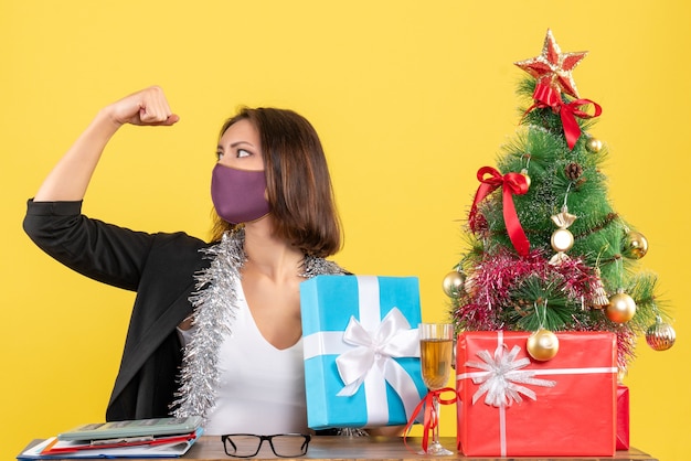 Weihnachtsstimmung mit stolzer schöner Dame im Anzug mit medizinischer Maske und Haltegeschenk im Büro auf Gelb