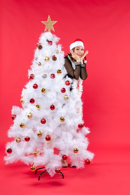 Weihnachtsstimmung mit süßer junger schöner Dame in einem schwarzen Kleid mit Weihnachtsmannhut, der sich hinter Neujahrsbaum versteckt