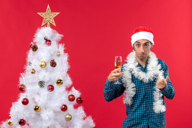 Weihnachtsstimmung mit traurigem jungem Mann mit Weihnachtsmannhut in einem blauen gestreiften Hemd, das ein Glas Wein nahe Weihnachtsbaum hält