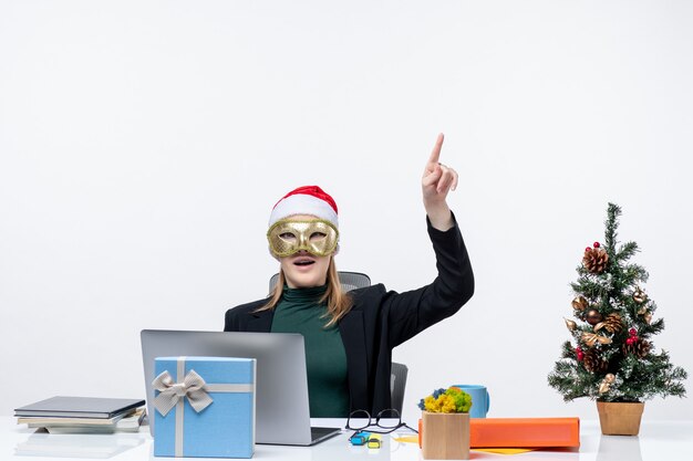 Weihnachtsstimmung mit überraschter junger Frau mit Weihnachtsmannhut und tragender Maske, die an einem Tisch sitzt, der oben zeigt, indem Fragen auf einem weißen Hintergrund stellt